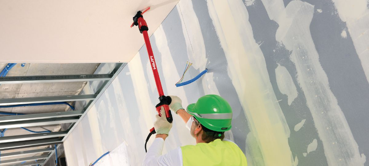 L084866 - Tanawa Construction worker in a green hard hat and high‑visibility vest mounts drywall ceiling using a red pole tool, with exposed wires nearby.